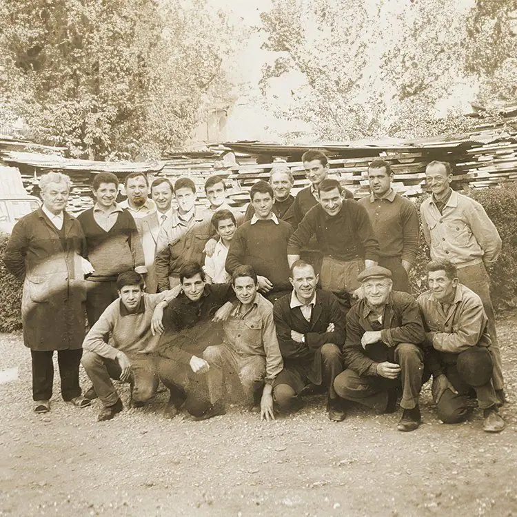 Fotografia d'epoca: il gruppo degli intagliatori della scuola, in posa all'aperto davanti a una catasta di legname
