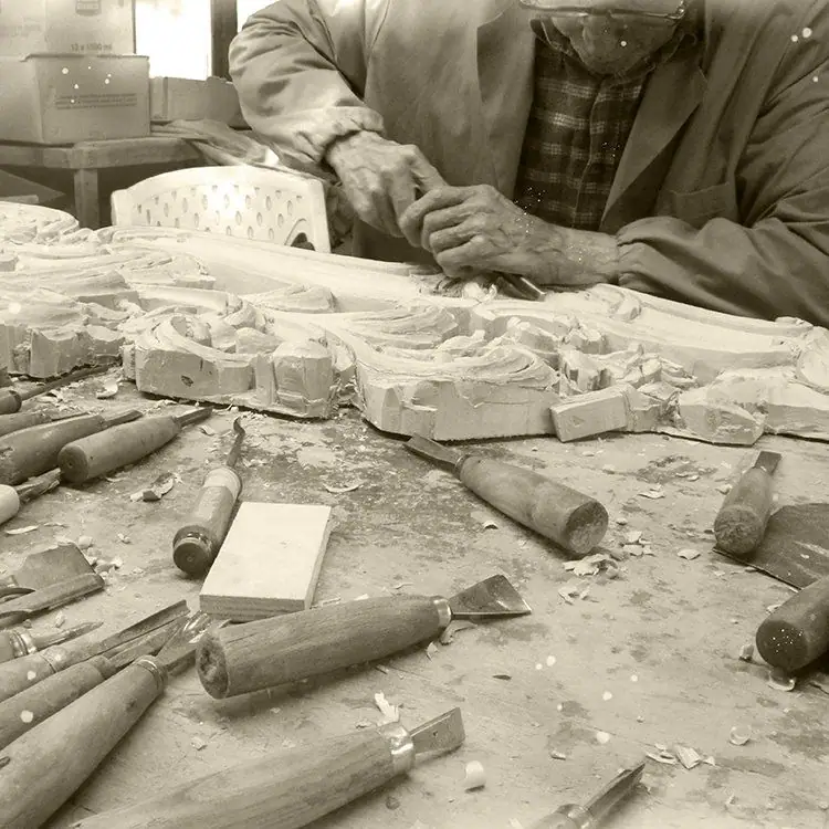 Fotografia in bianco e nero: le mani di un intagliatore al banco, fra sgorbie e pezzi di legno scolpito
